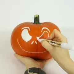 Ręce malują białą farbą wesołą buźkę na pomarańczowej dyni. Powstaje Halloweenowa dekoracja.