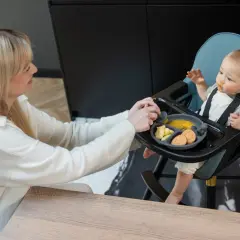 Mama karmi dziecko w krzesełku do karmienia, które nie siedzi samodzielnie. Na talerzyku jedzenie i ciasteczka.