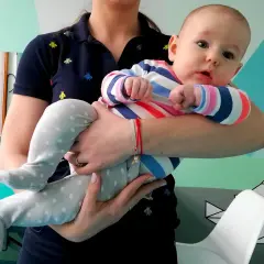 Malutka amfibia niemowlę w kolorowym pajacyku, trzymana przez opiekunkę, wygląda ciekawie na tle geometrycznych wzorów.