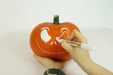 Ręce malują białą farbą wesołą buźkę na pomarańczowej dyni. Powstaje Halloweenowa dekoracja.