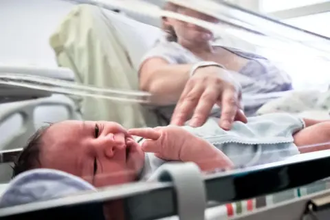 Noworodek, dziecko urodzone w 36 tygodniu, leży w inkubatorze. Mama delikatnie dotyka jego paluszka.