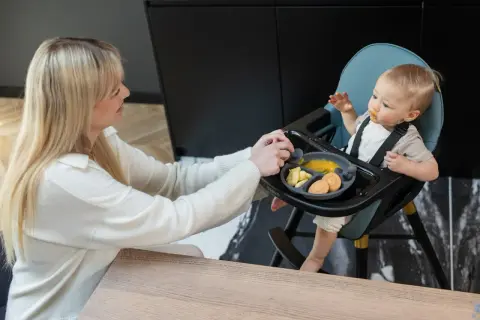 Mama karmi dziecko w krzesełku do karmienia, które nie siedzi samodzielnie. Na talerzyku jedzenie i ciasteczka.